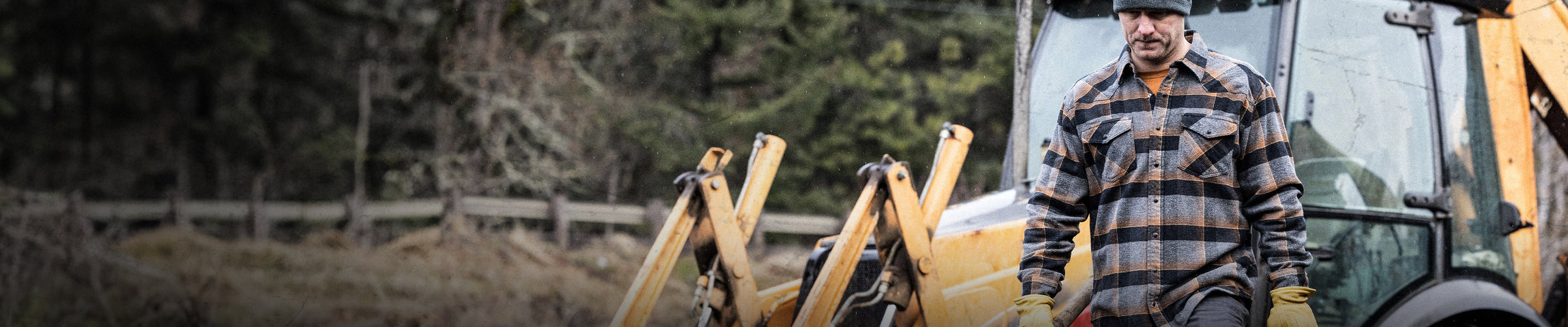 man in flannel shirt and gloves walking in front of heavy machinery