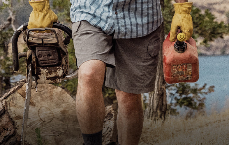 man in pants and shorts holding a gas can and chainsaw walking in front of a lake