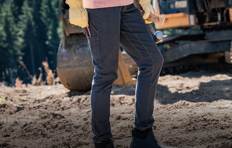 woman in gloves and pants standing in front of forest clearing with a large construction vehicle behind